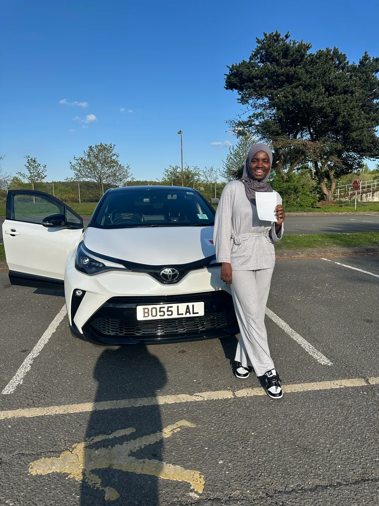 Zainab in a grey outfit and headscarf holding a faded pass certificate beside the training car.